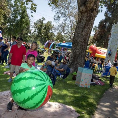 wide shot of picnic activities, families sitting together, kid playing with large inflatable watermelon, rock climbing wall and inflatable castle in background